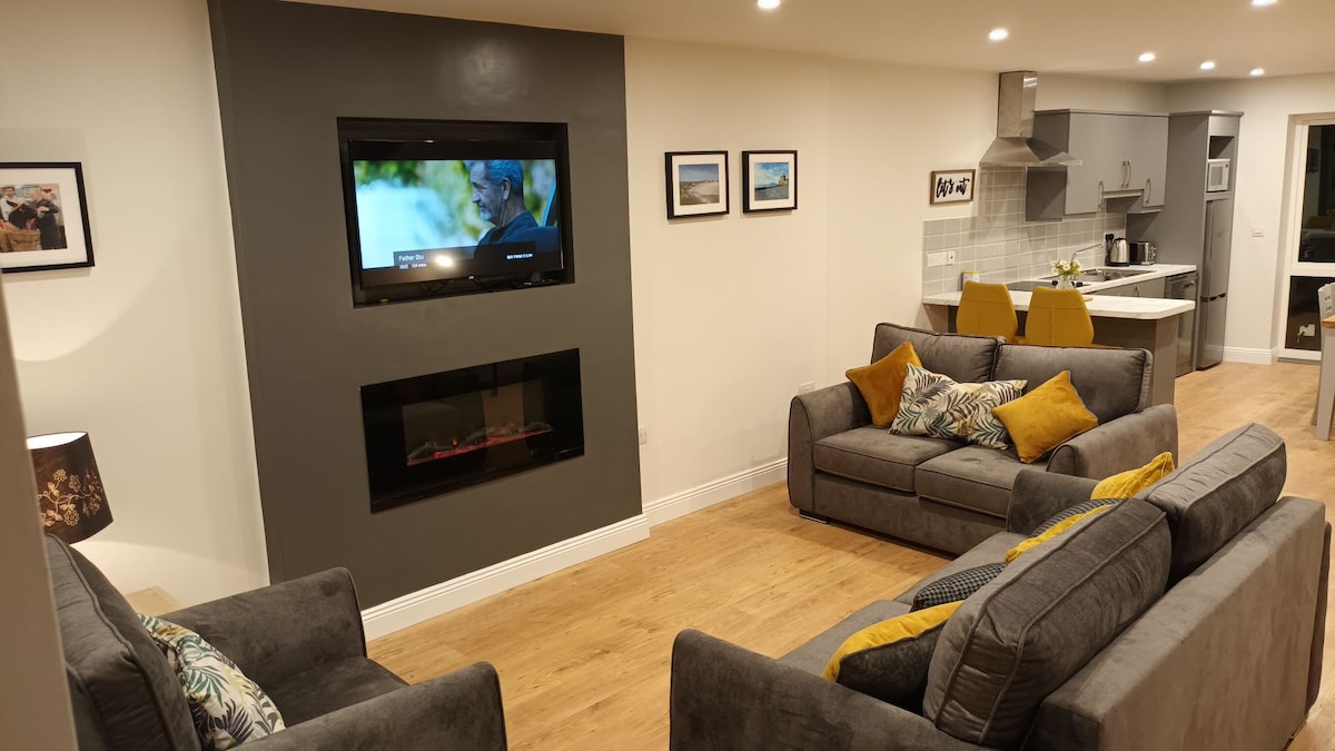 A modern living area features two gray sofas with colorful accent pillows arranged around a wall-mounted television. A decorative electric fireplace is positioned beneath the TV, adding a cozy touch. Light wood flooring complements the interior, and a glimpse of the kitchen area is visible in the background.