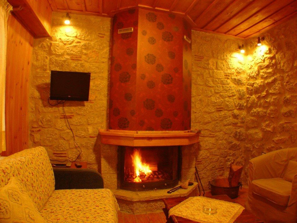 A cozy seating area features a stone fireplace, with a warm glow emanating from the flames. A flat-screen TV is mounted on the wall near the fireplace, and soft lighting complements the wooden ceiling and textured walls.