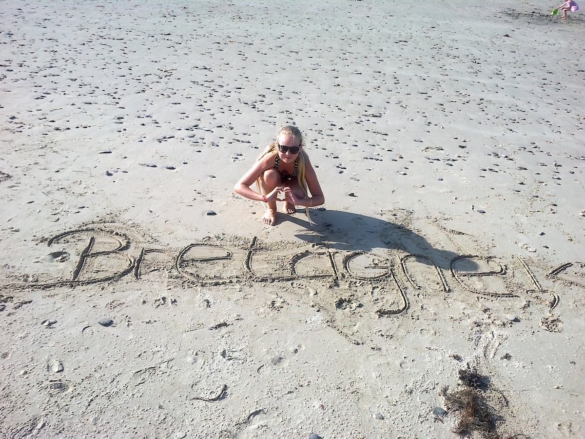 A sandy beach with a child kneeling beside the words 'Bretagne' written in the sand. Surrounding footprints are visible, indicating a lively beach atmosphere. The sea can be glimpsed in the background, enhancing the coastal setting.