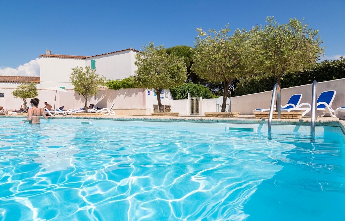 A clear blue pool is surrounded by sun loungers and olive trees, creating a serene outdoor setting. Sunlight reflects off the water's surface, while individuals relax nearby, enhancing the inviting atmosphere for leisurely afternoons.