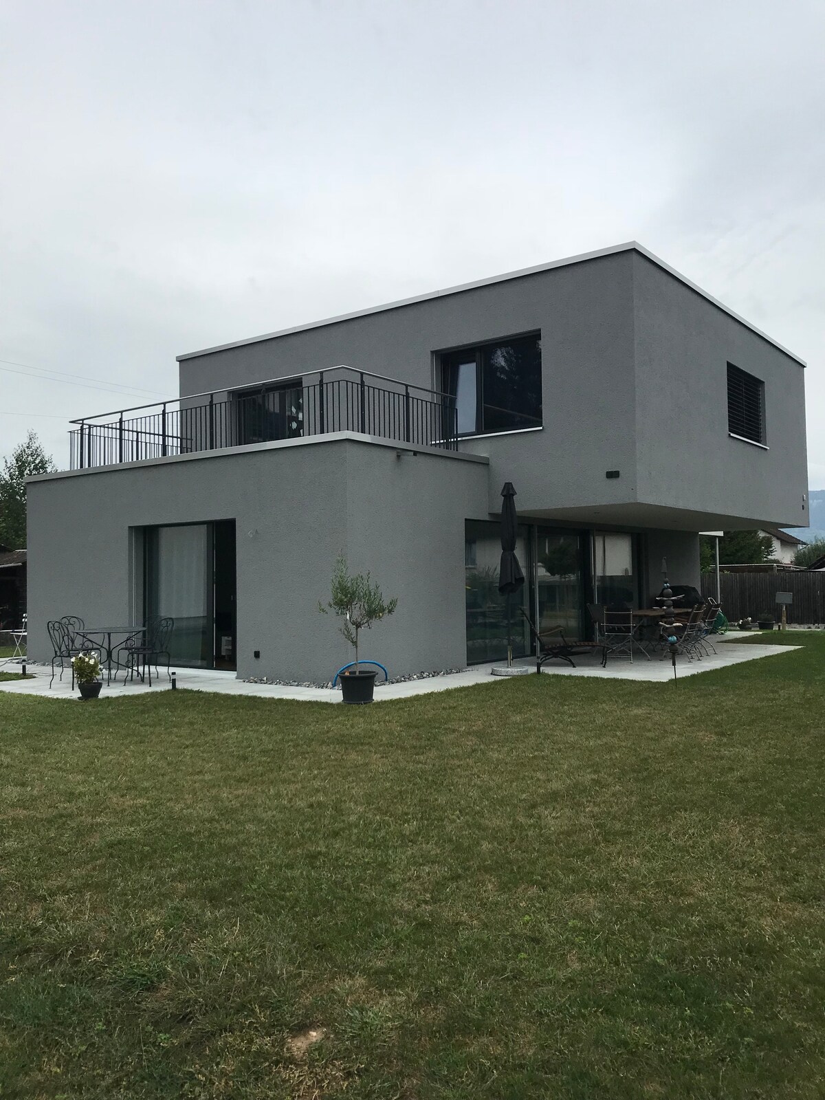 A modern two-story house is depicted with a sleek gray exterior. Large glass doors provide access to the outdoor area, where a small garden and seating arrangement are visible. A balcony extends from the upper floor, allowing for outdoor relaxation.