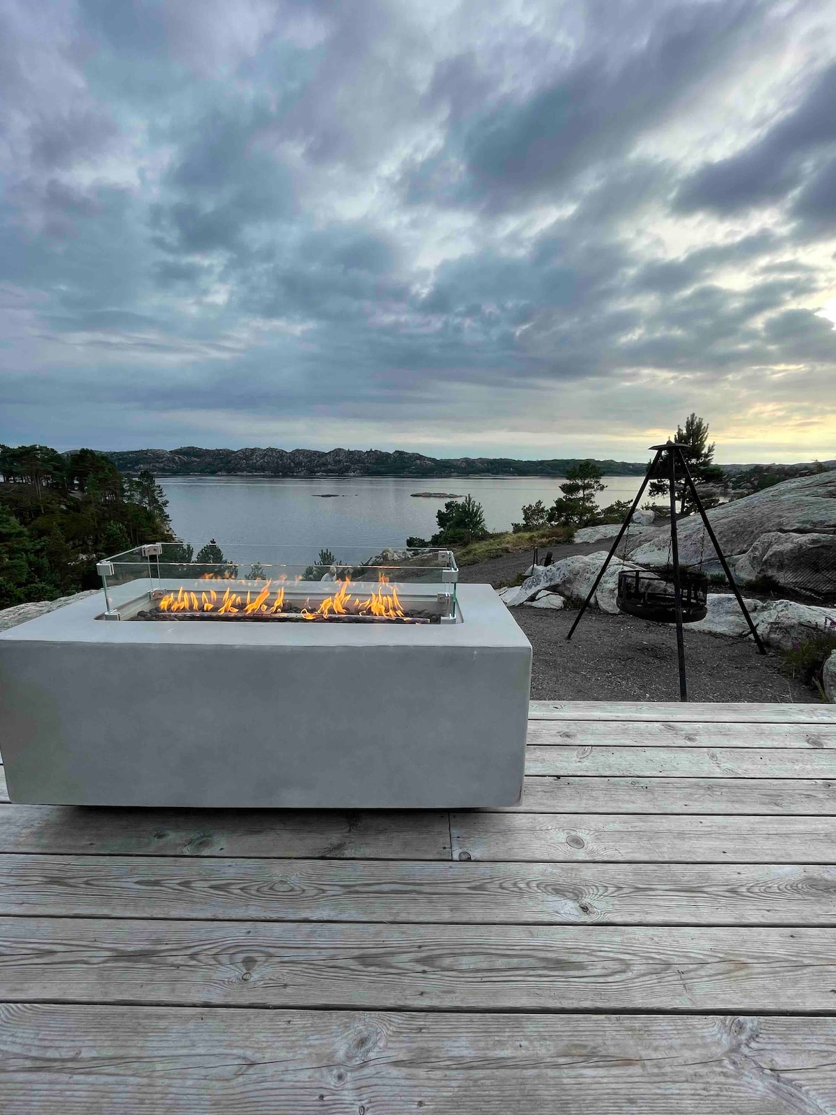 A modern fire pit is positioned on a wooden deck, offering views of the surrounding landscape and water. Flames are visible within a glass enclosure, under a cloudy sky that hints at sunset. A tripod stands nearby, complementing the serene atmosphere.