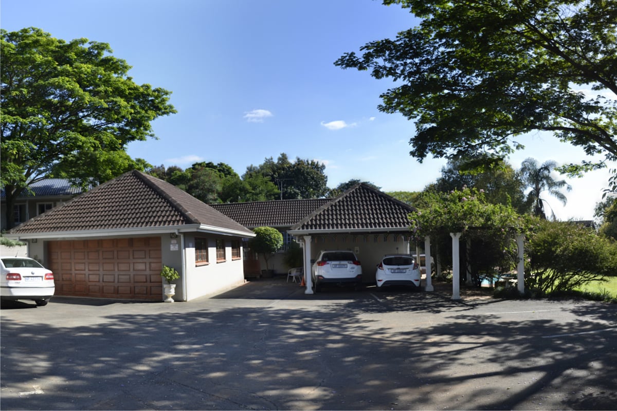 The exterior of the property features a well-kept garden with several trees, providing shade. Two cars are parked in designated spots, and a garage is visible on the left side. The structure displays a two-tone exterior, complemented by a tiled roof and a cozy porch area.