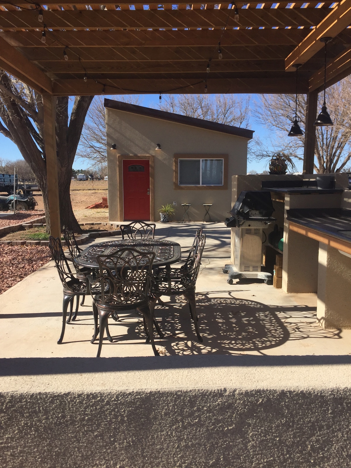 An outdoor patio area is highlighted, featuring a metal dining set with multiple chairs surrounding a round table. A grill is positioned nearby under a wooden pergola, casting interesting shadows on the ground. The guest house entrance is visible in the background, showcasing a bright red door.