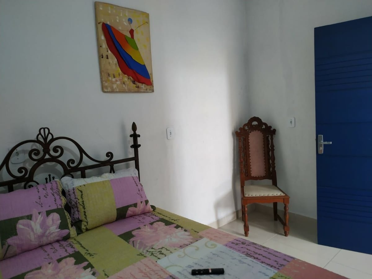 A comfortable sleeping area is presented with a patterned bedspread featuring floral designs. An iron bed frame supports two pillows. A decorative wall art piece adds color above the bed. A wooden chair is placed nearby, and a blue door leads to another space.