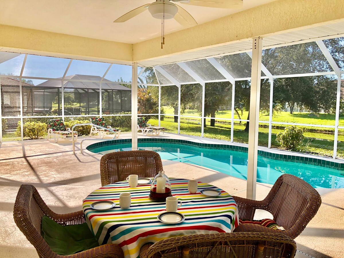 A dining area features a round table adorned with a colorful striped tablecloth, surrounded by wicker chairs. A screened-in patio overlooks the heated pool, with green grass and trees beyond, providing a tranquil outdoor setting.