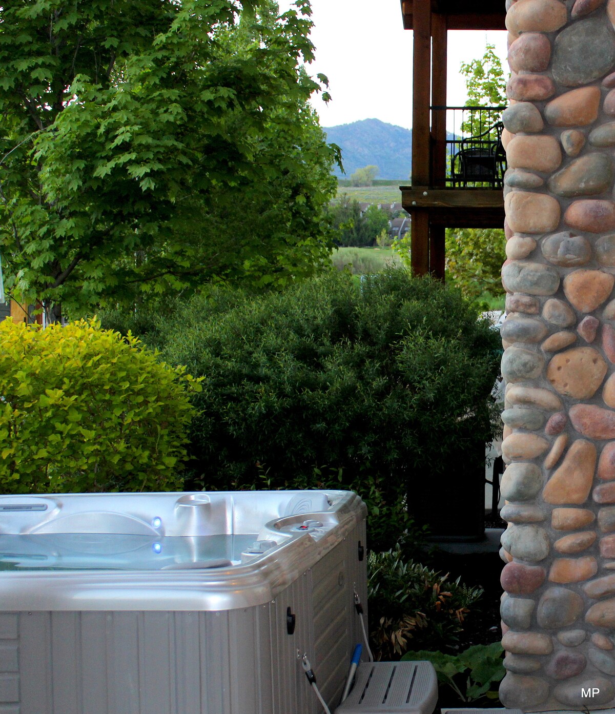 A private hot tub is situated near a stone wall, surrounded by lush greenery and colorful bushes. The backdrop features distant hills and trees, creating a serene atmosphere. The area is designed for relaxation and is easily accessible from the studio apartment.