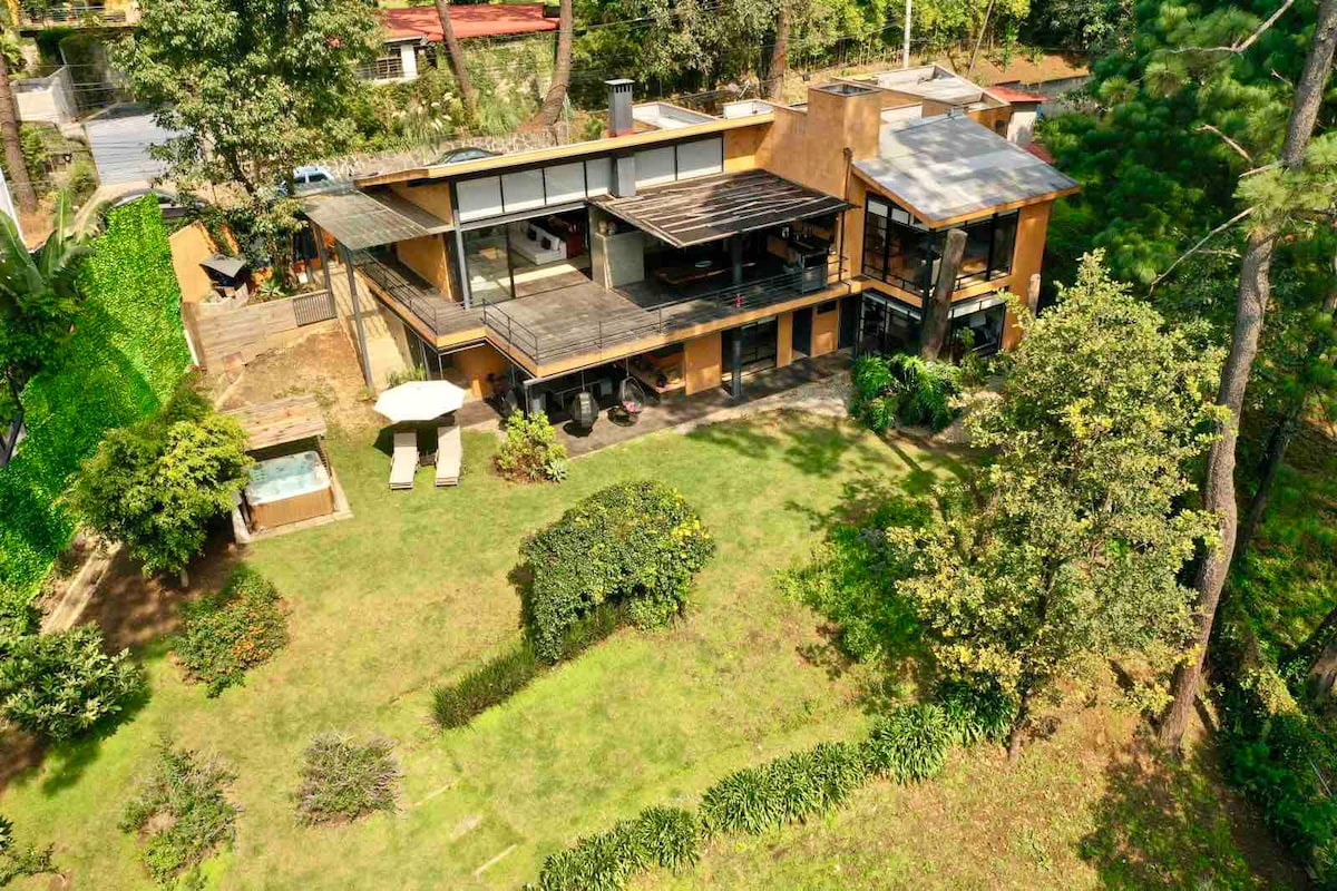An aerial view of a modern house is presented, surrounded by lush greenery. A well-maintained garden features a jacuzzi area, two lounge chairs, and a spacious lawn. The house design showcases large windows and multiple outdoor spaces, highlighting the harmonious integration with nature.