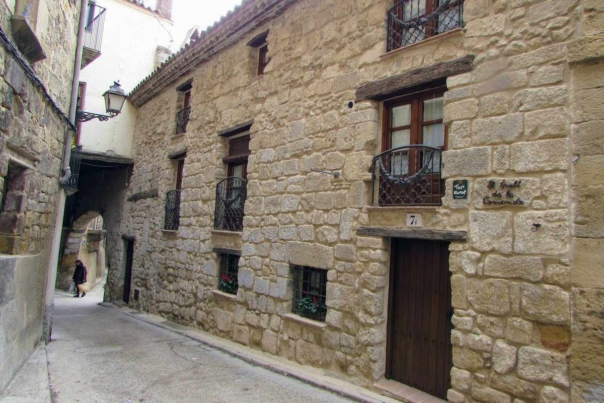 A historic stone building is seen along a narrow, cobblestone street, featuring wrought iron balconies with flower boxes. The entrance is adorned with wooden doors and a plaque displaying the house name, while muted walls reflect the architectural style of the local area.