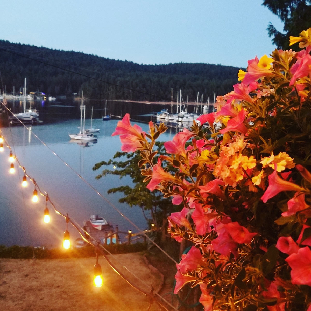 A vibrant display of pink flowers is framed in the foreground, with twinkling string lights illuminating the scene. In the background, calm waters reflect the silhouettes of sailboats and lush green hills under a twilight sky.