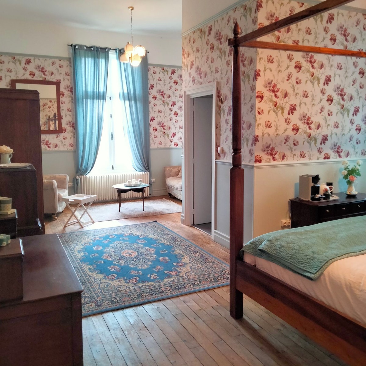 The spacious room features floral wallpaper and wooden flooring. A four-poster bed sits beside an antique dresser. A seating area with a light-colored couch and small table is present, along with a blue area rug that adds a touch of color.