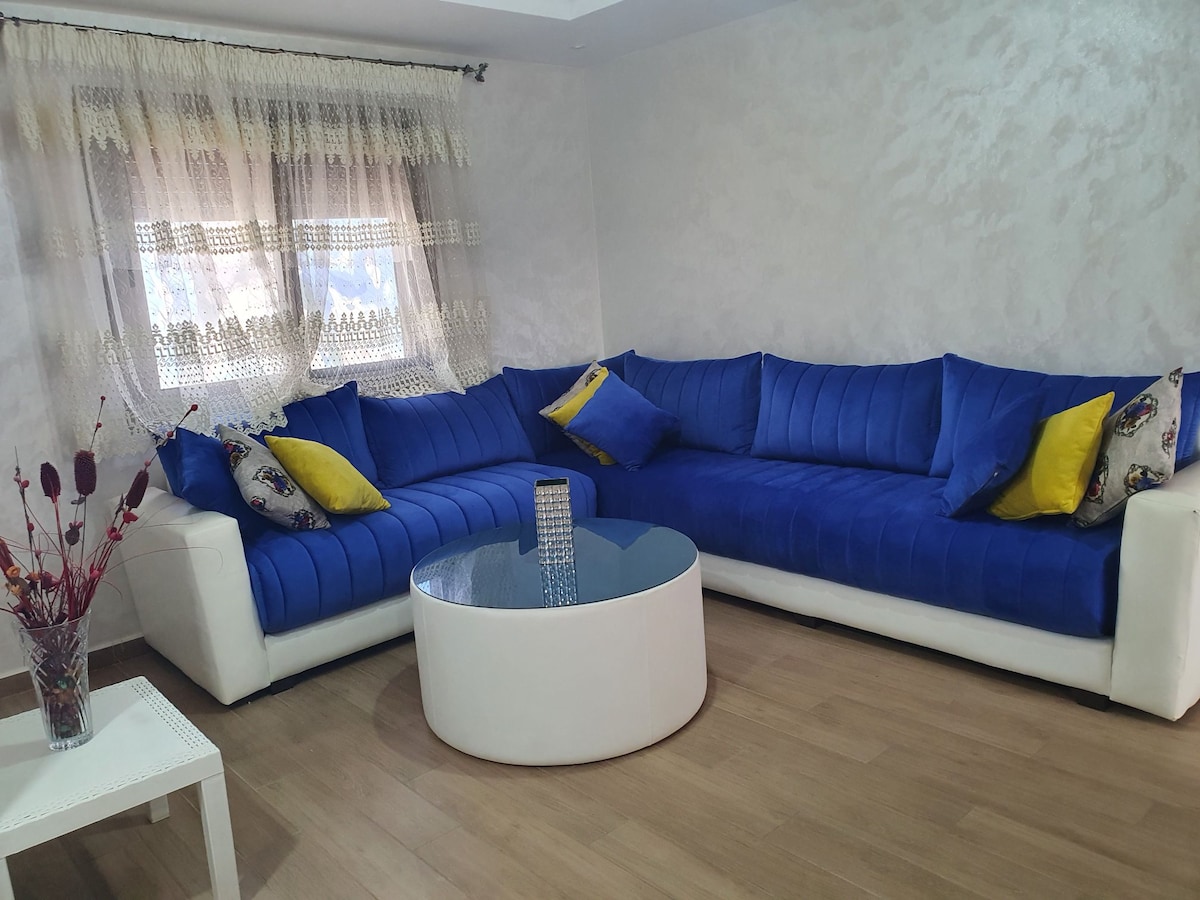A spacious living area features a large L-shaped sectional sofa in vibrant blue, adorned with yellow and patterned cushions. A round glass coffee table is centered in front, and soft natural light filters through lace-curtained windows, creating a comfortable gathering space.