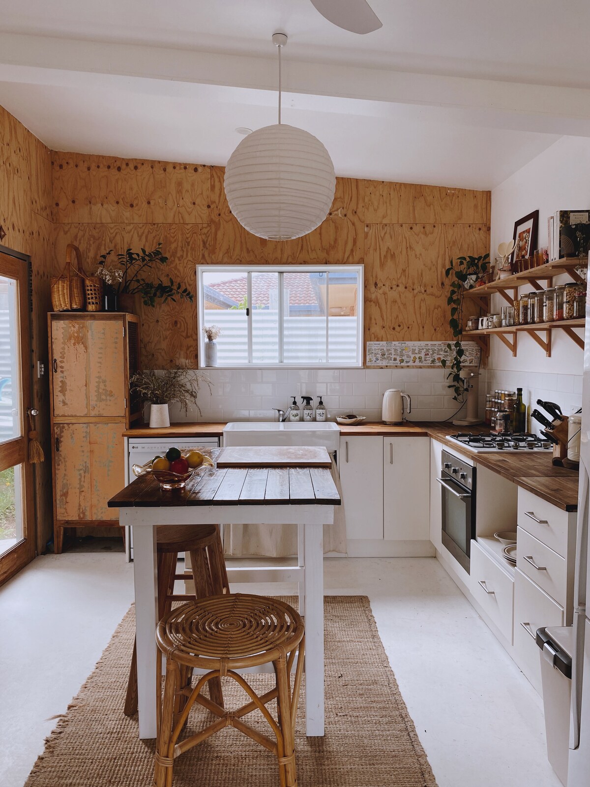 The kitchen area features white cabinetry paired with wood paneling. A simple wooden dining table is set at the center, accompanied by a wicker chair. Bright natural light flows in through a window, showcasing a gas cooktop beside an assortment of dishes and kitchenware on shelves.