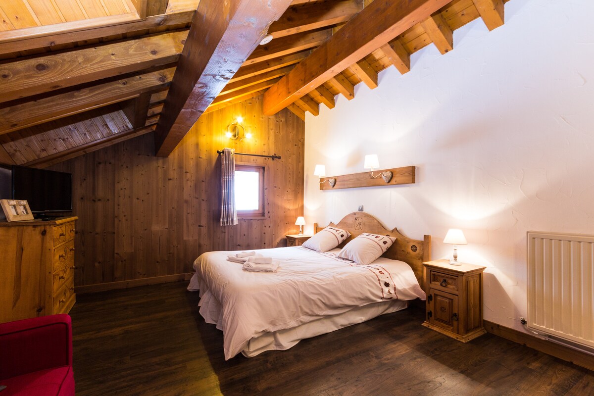 A spacious room features a large double bed, complemented by bedside tables with soft lighting. The wooden ceiling beams add warmth, while wooden paneling creates a cozy ambiance. A small window allows natural light to illuminate the area.