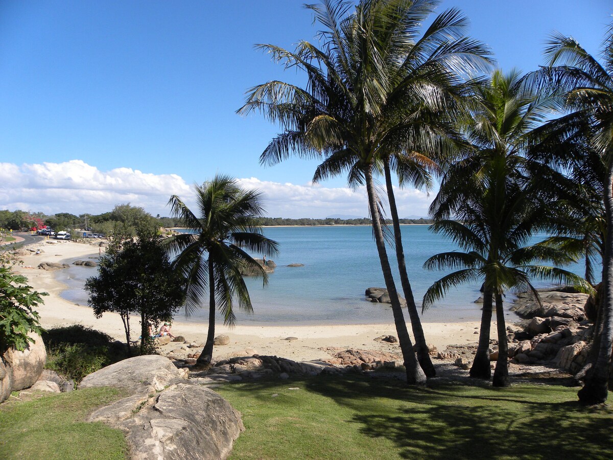 A scenic view of a tranquil beach surrounded by palm trees. The calm water reflects the clear blue sky, while a sandy shore meets smooth rocks. Lush green grass leads down to the water's edge, offering a serene atmosphere for relaxation.