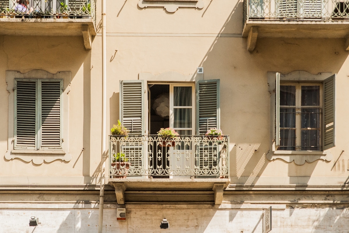 The image features a building facade with light-colored walls and decorative accents. Two balconies are adorned with flower pots, and wooden shutters are partially open, revealing a glimpse of the interior. A decorative wrought iron railing complements the overall charm.