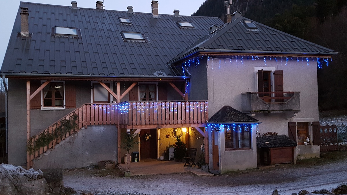 An old farm building, renovated, with a sloped gray roof and wood accents. Decorative blue lights adorn the exterior, highlighting the entrance and balcony. A peaceful, mountainous landscape is visible in the background, adding a serene touch to the setting.