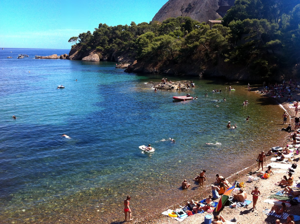 A sandy beach is visible, bordered by clear blue waters. Sunbathers are spread across the shoreline, and swimmers can be seen enjoying the water. Small boats are anchored in the distance, surrounded by lush greenery that frames the beach.