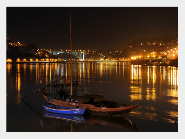 A tranquil scene is captured at night, featuring traditional boats moored on calm waters. Illuminated reflections from waterfront buildings and lights create a warm glow, enhancing the serene atmosphere of the riverside.