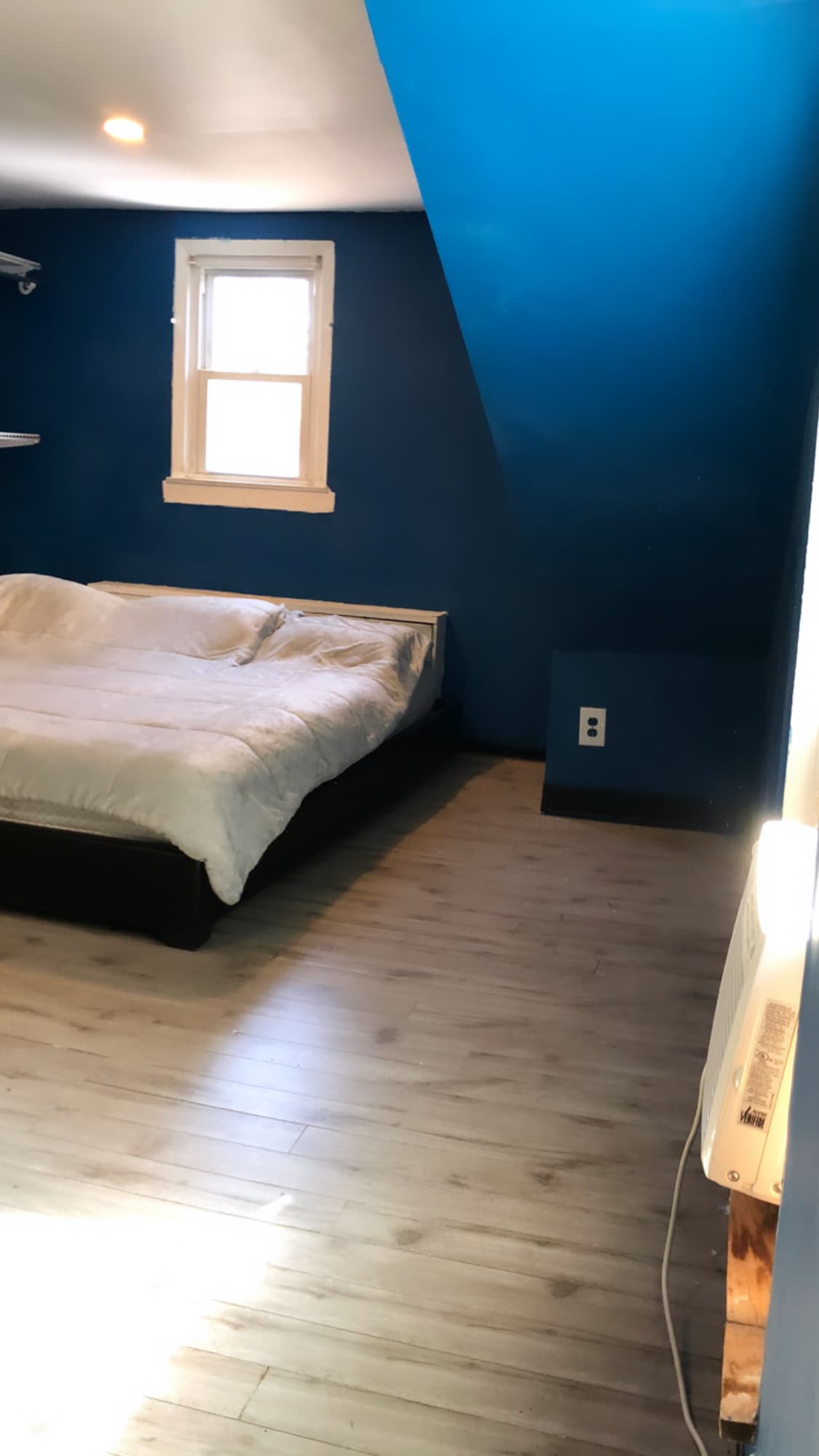 The spacious room features a California KING size bed with white bedding, situated against a striking blue wall. Shelving units are mounted above the bed, and natural light enters through a nearby window, enhancing the calming atmosphere of the space.