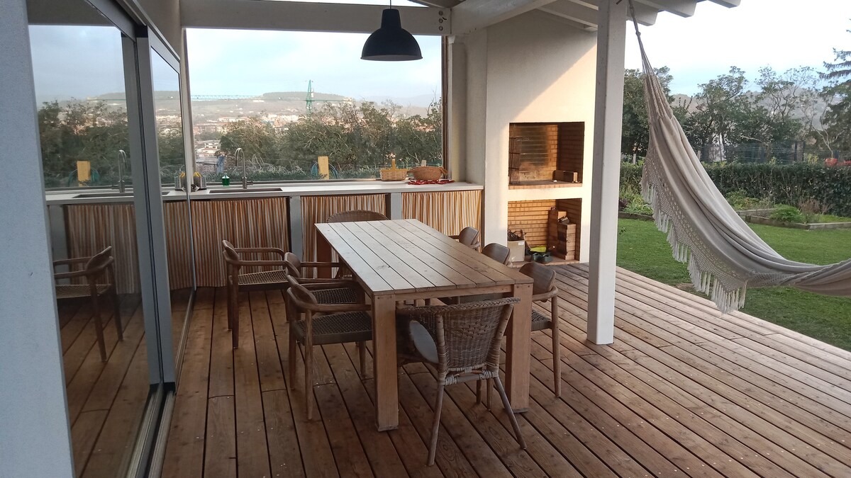 An outdoor porch features a wooden dining table surrounded by six woven chairs. A hammock is hung to one side, providing a relaxed seating option. A built-in outdoor barbecue is visible against a wall, and large glass doors open to the interior space.