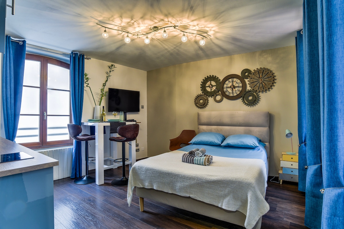 A cozy studio space features a spacious bed with light bedding. Unique wall decor includes decorative gears. An inviting work area is present with two stools and a television mounted on the wall. Large windows with blue drapes allow natural light to brighten the room.
