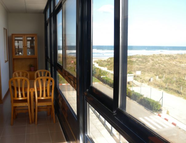 A dining area is visible through large glass windows, featuring a wooden table surrounded by four chairs. The view beyond includes a sandy beach and ocean waves, creating a bright and airy setting.