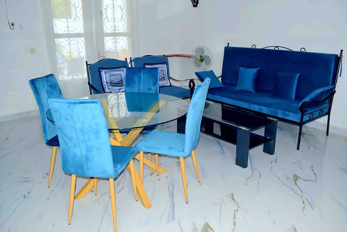 A dining area features a circular glass table surrounded by six blue velvet chairs. A sleek black coffee table is positioned nearby, complementing a sofa adorned with matching blue cushions. Natural light illuminates the space through large windows with decorative ironwork.