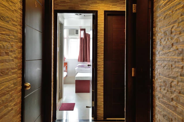 A corridor with textured stone walls leads to an open door revealing a brightly lit guest room. Light filters through sheer red curtains, highlighting the simple furnishings within. The flooring features a polished finish, enhancing the space's clean and organized appearance.