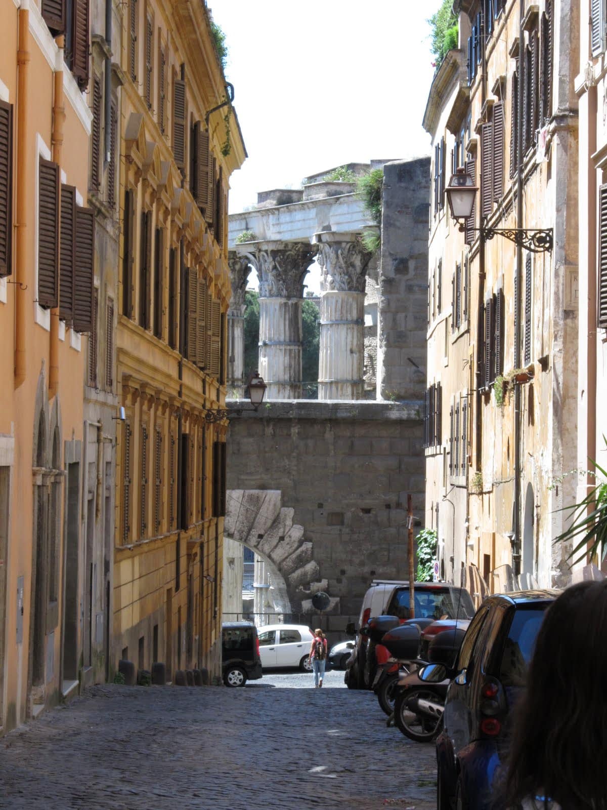 A narrow street lined with historic buildings showcases vibrant yellow and warm brown facades. At the end of the street, ancient columns are visible, hinting at the storied past of the area. Cars are parked along the cobblestone path, adding to the urban scene.