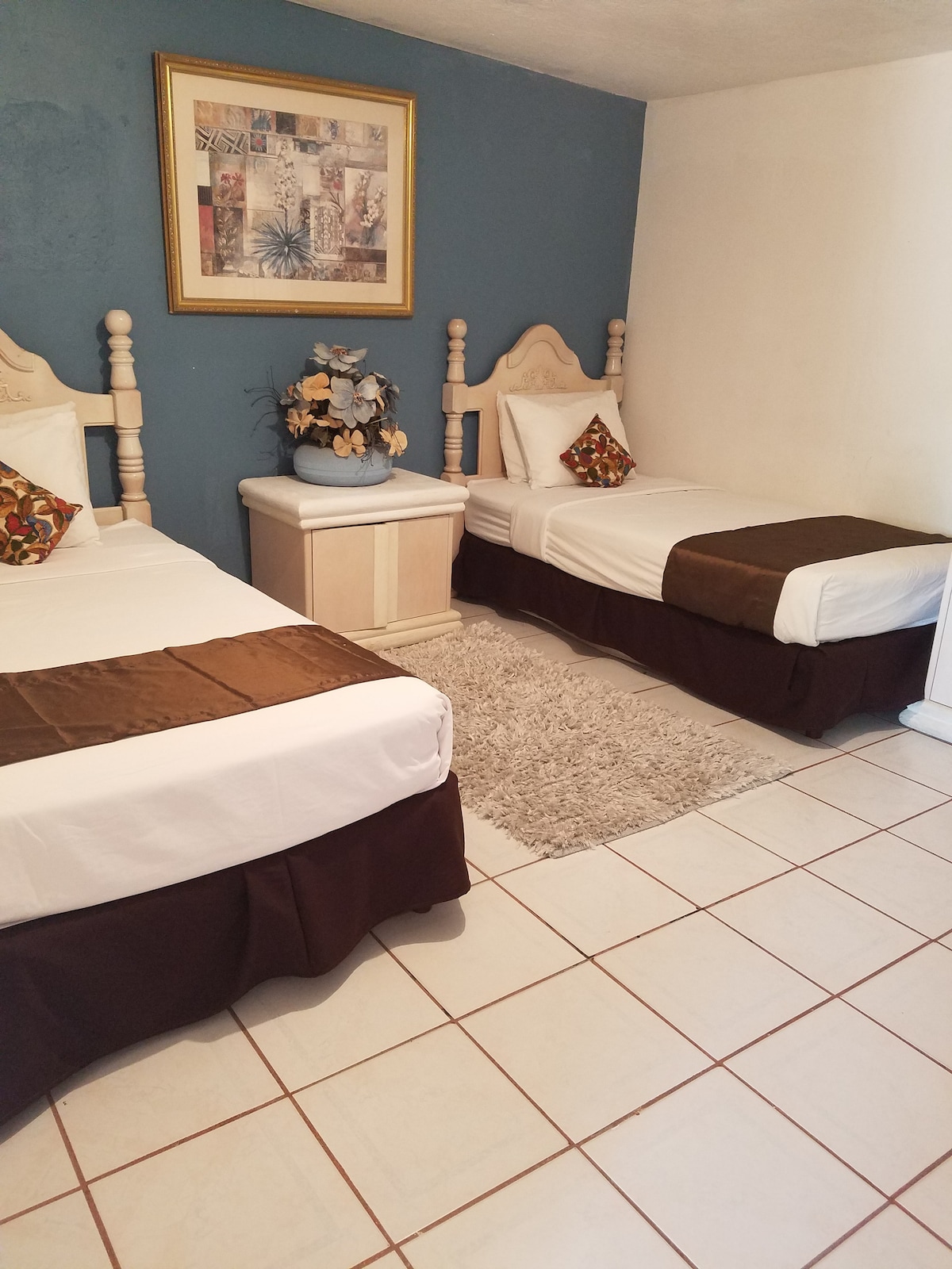 A cozy bedroom is presented with two neatly made twin beds, each styled with decorative pillows. A small nightstand is positioned between the beds, adorned with a vase of flowers. A plush rug covers the tiled floor, adding comfort to the space.