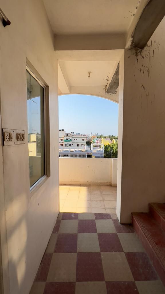 A corridor is depicted leading towards a balcony. Checkerboard tiles in red and white cover the floor. Natural light enters through a large window, illuminating the space. A wall-mounted electrical panel is visible next to the entrance, and stairs are seen leading down.