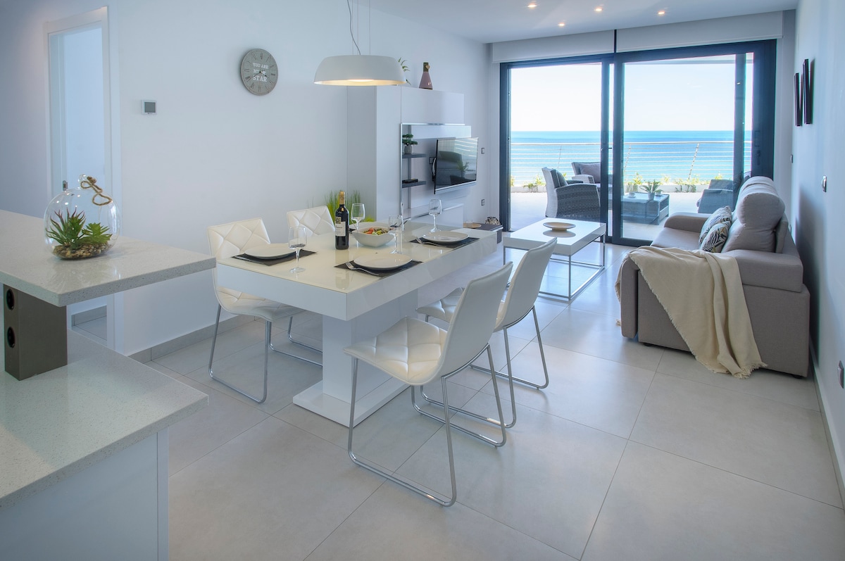 A modern dining area is equipped with a sleek white table and four stylish chairs, set against a backdrop of large glass doors that offer ocean views. A comfortable sofa faces the sea, creating a spacious ambiance filled with natural light.