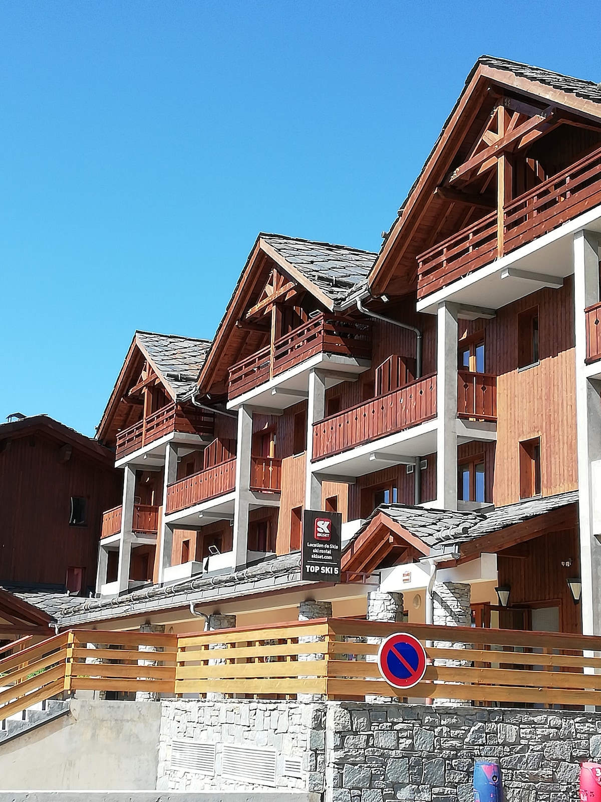 A multi-story building features wooden balconies with decorative railings, characteristic of alpine architecture. The facade showcases a blend of stone and timber, complemented by the bright blue sky above, creating a clean and inviting appearance.