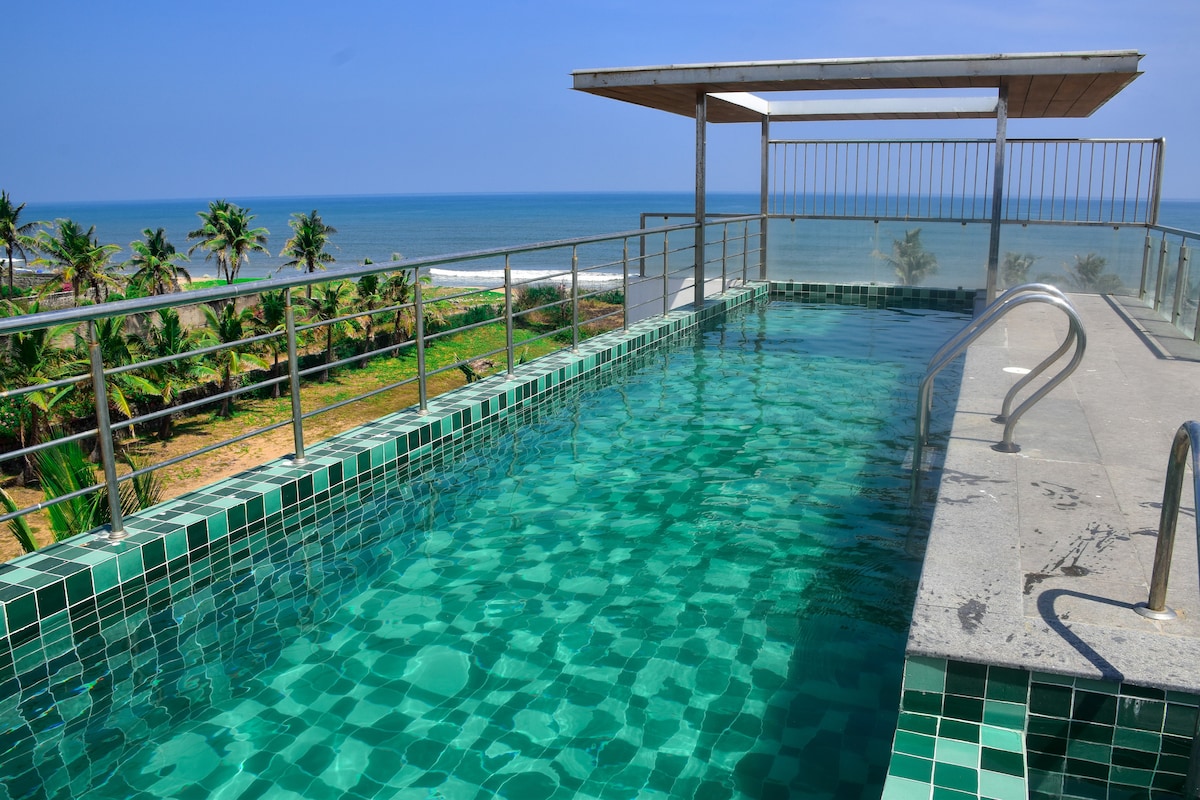 A rooftop swimming pool offers stunning views of the ocean, with clear turquoise water inviting relaxation. Sunlight dances on the surface, while surrounding palm trees sway gently in the breeze, complementing the serene coastal atmosphere.