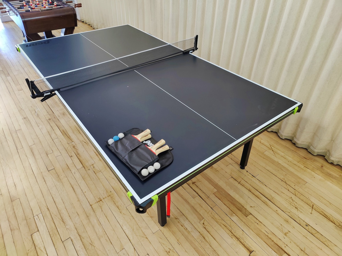 A ping pong table is set up in an arcade room, featuring a black surface with a white boundary. Paddles and balls are neatly arranged on a carrying case beside the table. Light wood flooring complements the space.