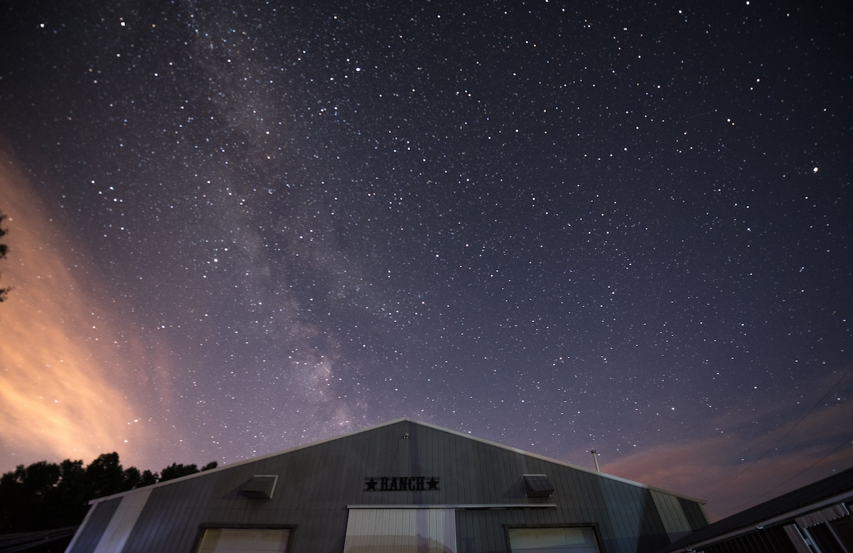A night sky filled with stars is captured above a vintage guest ranch. The Milky Way is visible, providing a backdrop against the structure's metallic exterior. The serene ambiance suggests a peaceful retreat in nature.