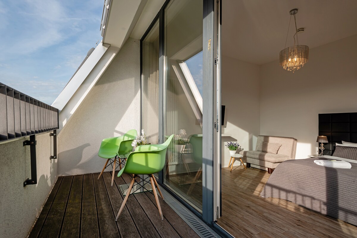 A balcony area features two green chairs beside a small table, complemented by a spacious indoor seating area visible behind glass doors. Natural light fills the room, accentuating the wood flooring and modern furnishings that include a cozy sofa and neatly arranged bed.