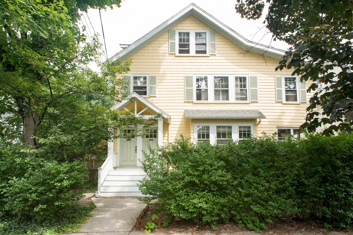 A charming two-story yellow house is framed by lush green shrubs and trees. The front porch features a covered entrance with a set of steps leading up to double doors. Large windows are visible on the upper floors, enhancing the light and airy appearance.