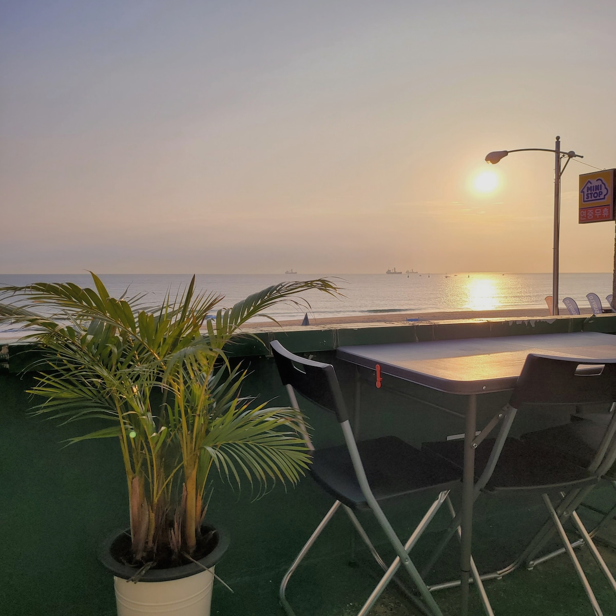 A potted plant is positioned next to a table and chair setup on the rooftop terrace. The sun sets over the ocean in the background, reflecting light on the water's surface. Silhouettes of ships are visible on the horizon, enhancing the coastal view.