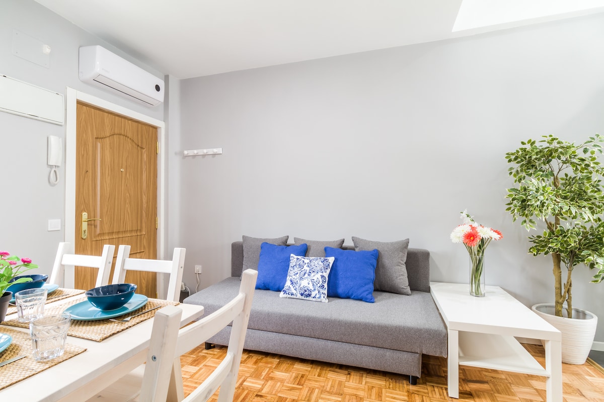 A light-filled living area features a gray sofa adorned with blue and white cushions. A small white table and a potted plant are visible, alongside a dining table set with blue dishware and glassware. A light wooden door leads to the entrance.