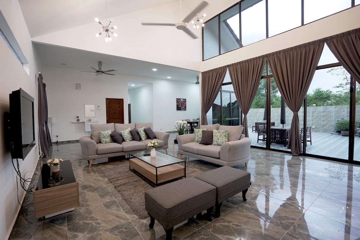 A spacious living area features high ceilings and large windows, allowing natural light to fill the room. Comfortable sofas are arranged around a low coffee table, and decorative cushions add subtle color. Dark curtains frame the windows, while an open view of the outdoor space is visible.