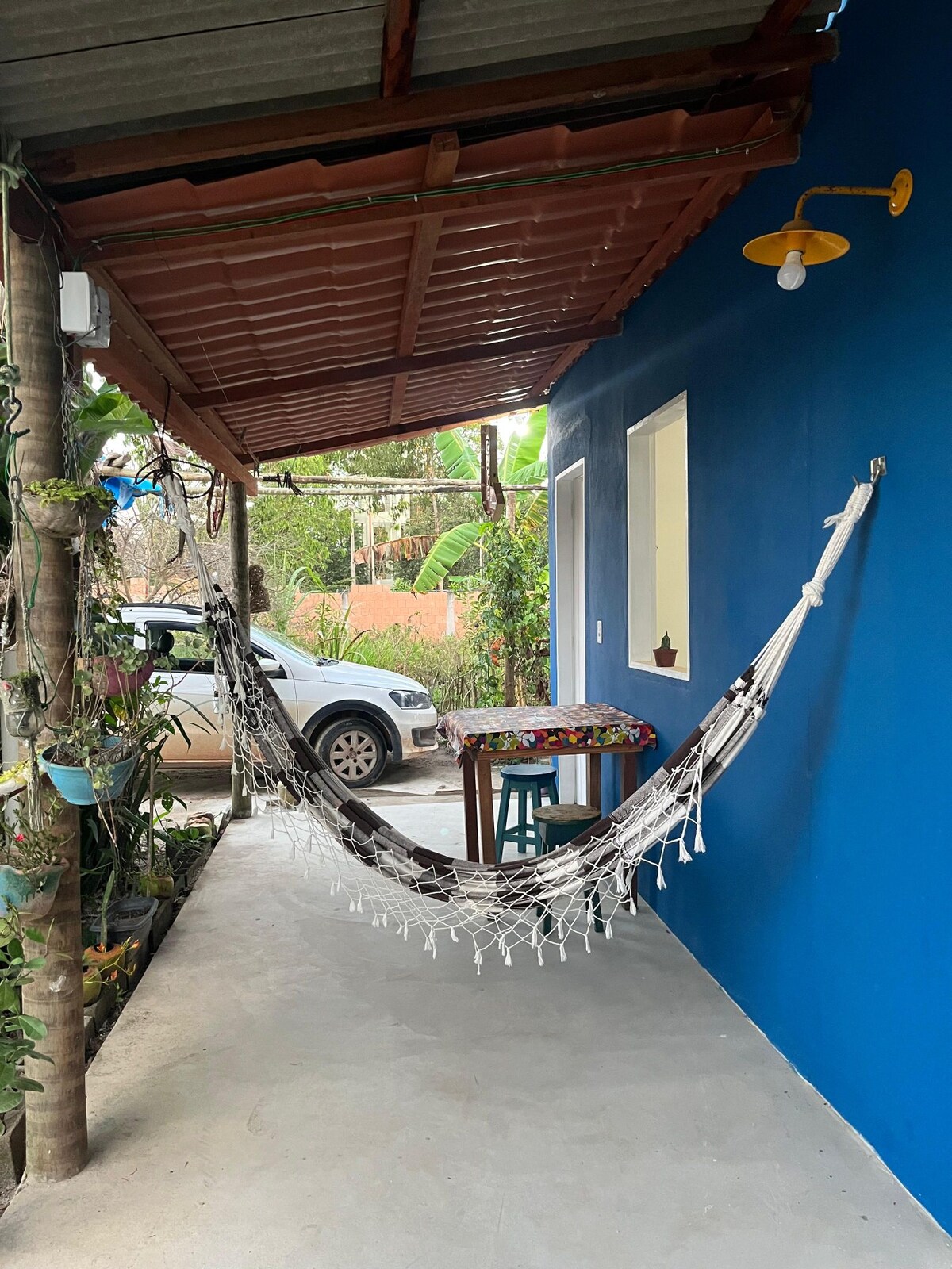 A shaded outdoor area features a hammock strung between the posts, inviting relaxation. A small table sits nearby, adorned with colorful decorations. Lush plants line the space, and a vehicle is parked just outside. The blue wall adds a vibrant touch to the setting.