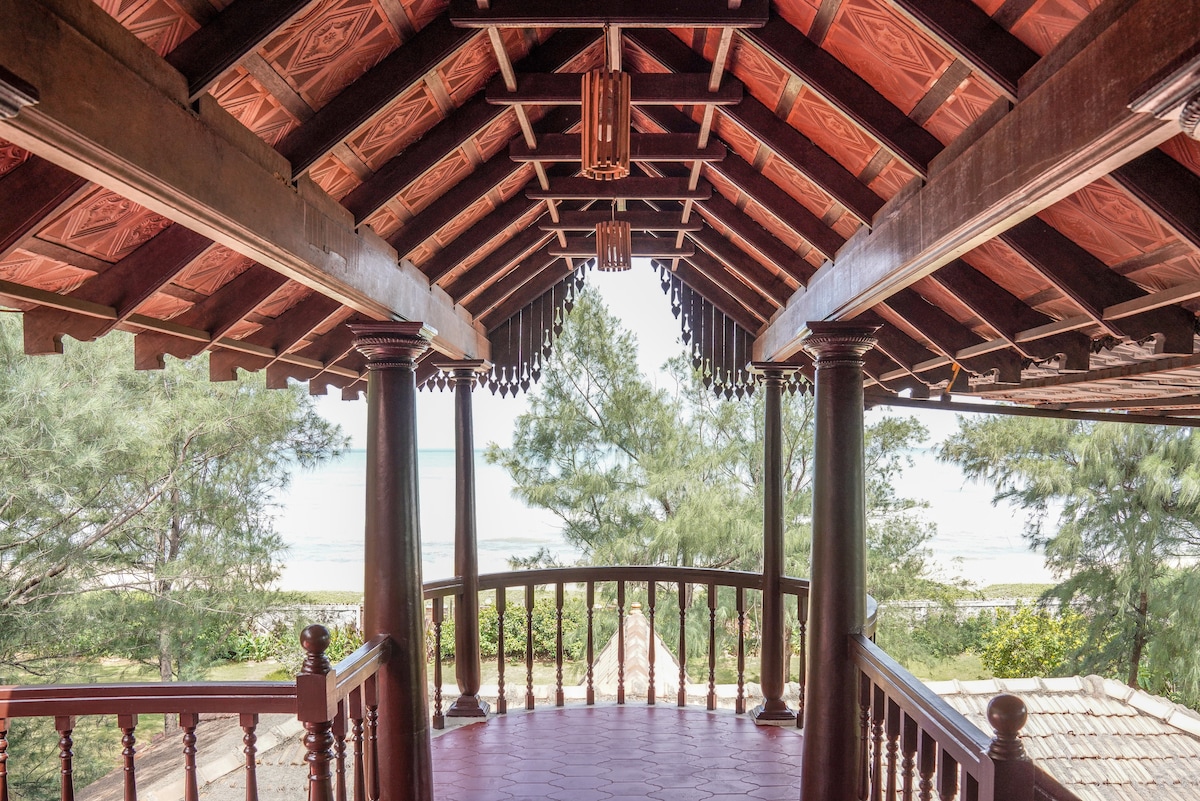 A spacious balcony with a high wooden ceiling, featuring ornate detailing and decorative light fixtures. The view reveals lush green trees and calm sea waves in the distance, inviting relaxation in a serene setting.