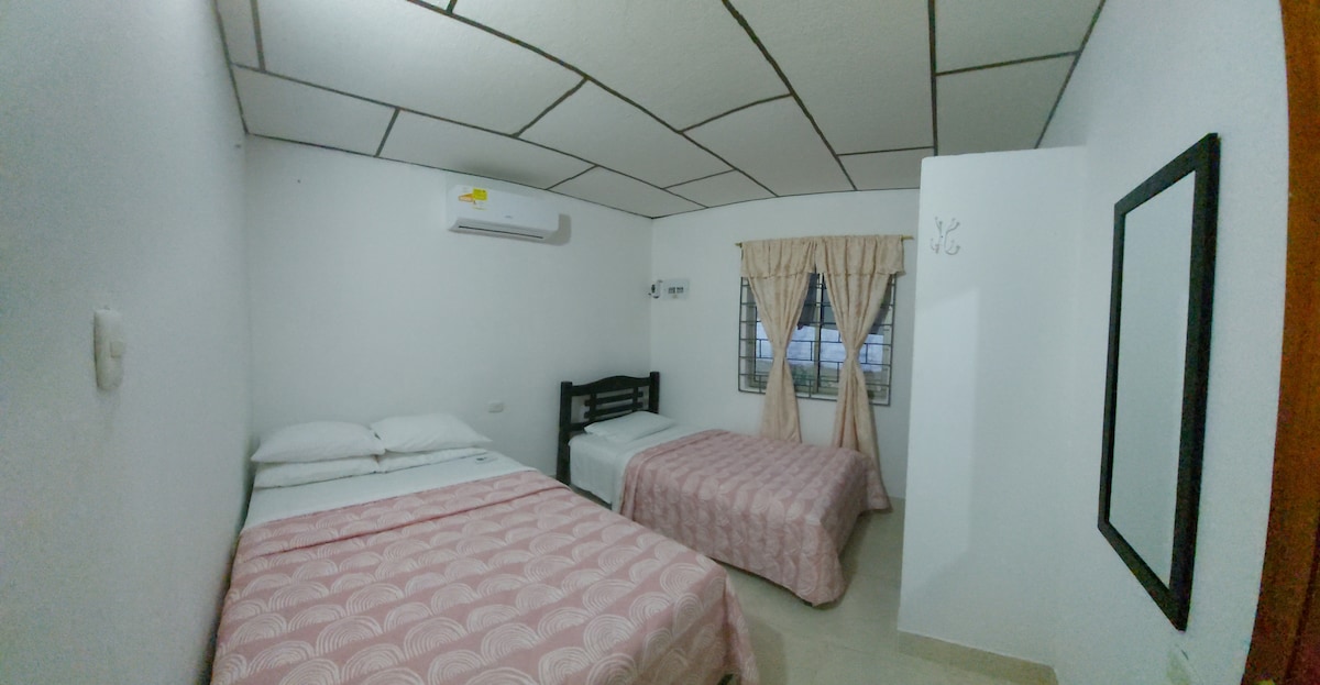 The room features two beds, one with a light pink patterned quilt and the other with a solid white cover. A window with sheer curtains allows natural light in, while an air conditioning unit is mounted above. A mirror and hooks are visible on the wall.