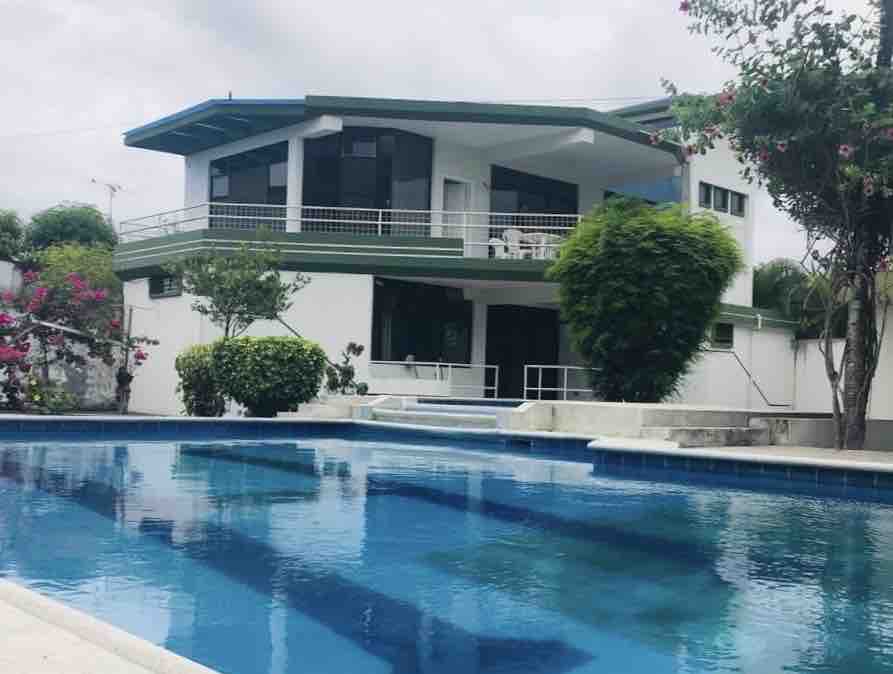 A contemporary house is set against a backdrop of lush greenery, featuring large windows that allow for ample natural light. A clear blue swimming pool reflects the surrounding landscape, creating a serene outdoor space. Vibrant bushes add color to the scene.