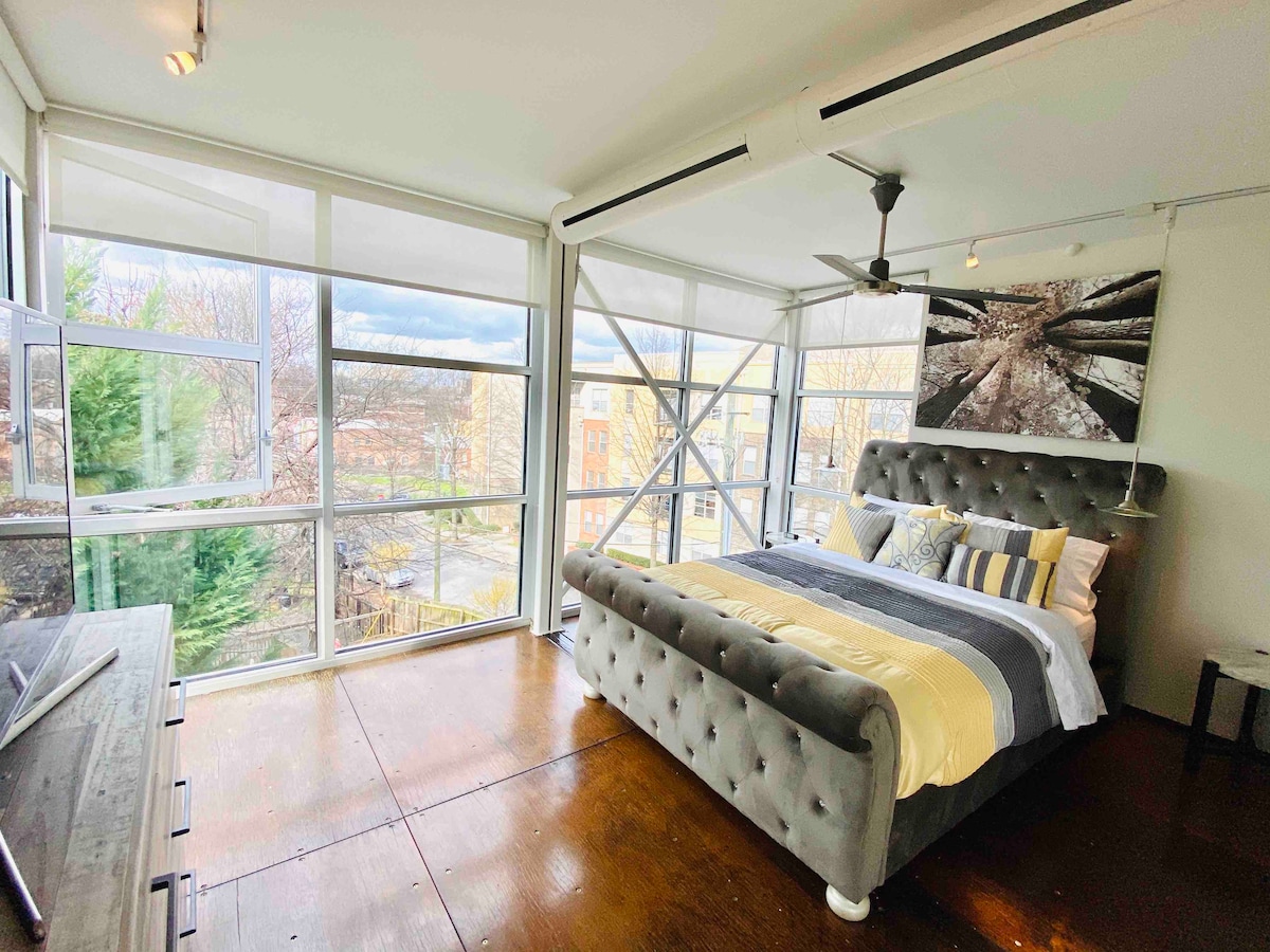 A spacious bedroom features a modern grey upholstered bed with striped bedding. Large glass windows provide ample natural light and offer views of the surrounding area. A ceiling fan is visible above, and a simple desk is positioned nearby, all set against a wooden floor.