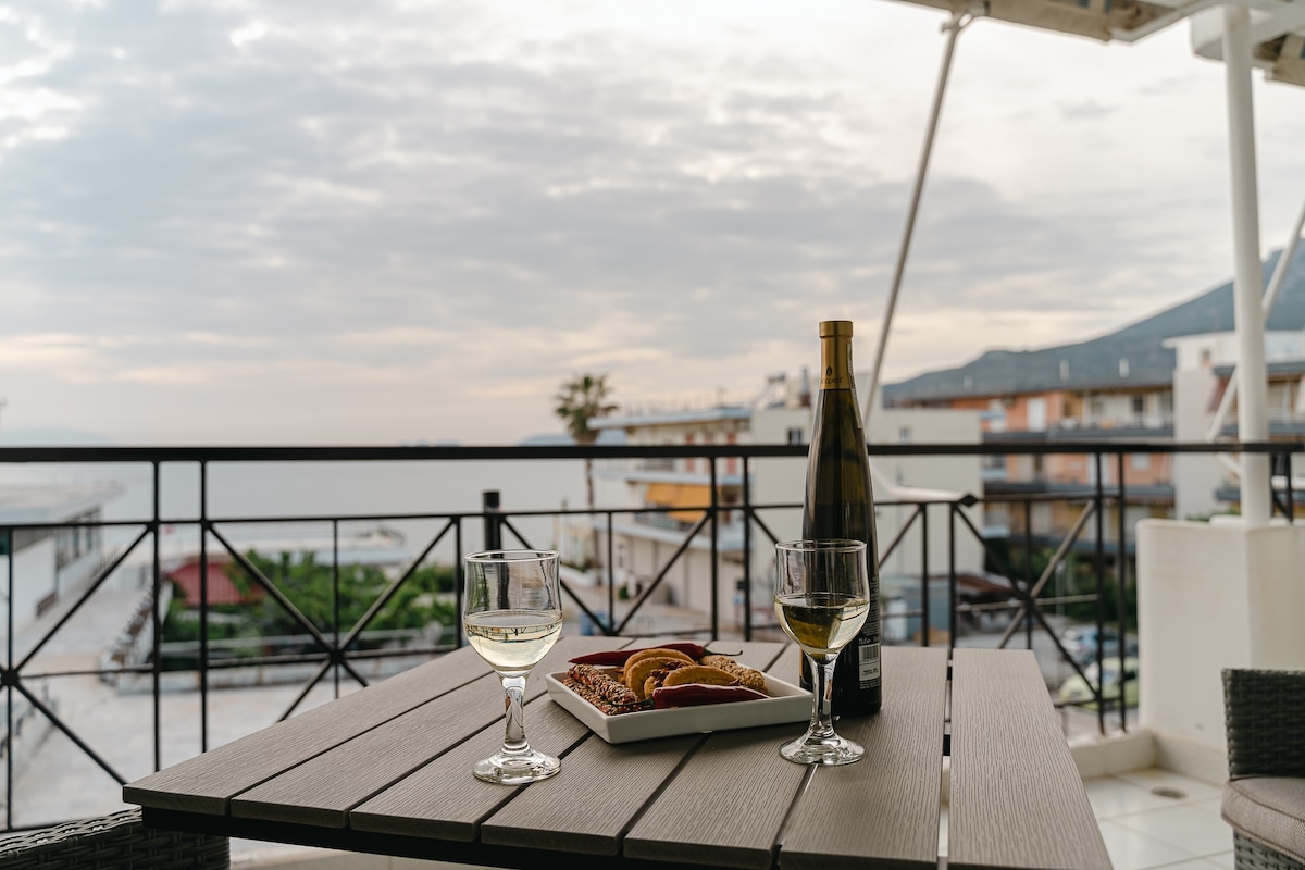 A balcony table is set for two, featuring two glasses of white wine alongside a plate of snacks. The serene sea view is visible in the background, with soft clouds overhead and buildings lining the waterfront.