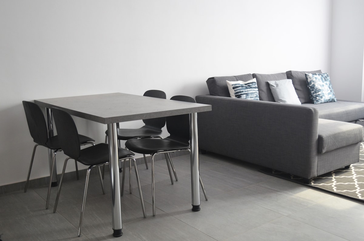 A modern dining area is visible, featuring a rectangular table surrounded by six black chairs. Adjacent to the dining area, a gray sofa is positioned, decorated with cushions in various shades of blue. A patterned rug lies underneath the furniture, enhancing the overall space.