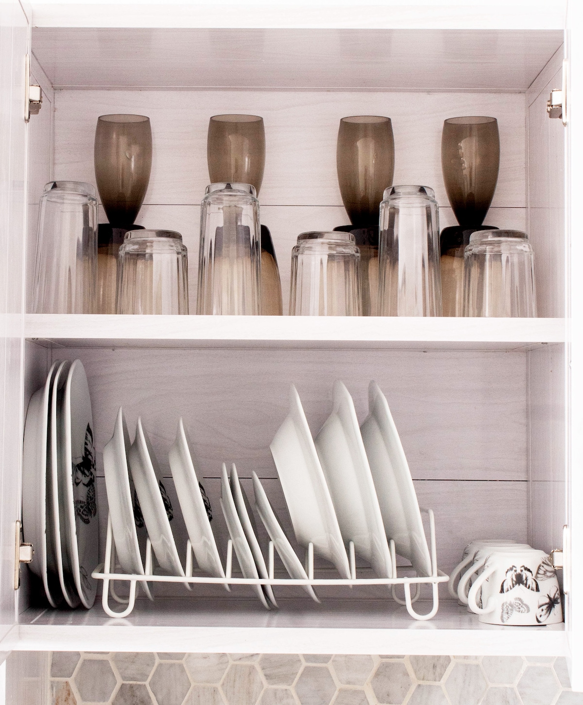 A neatly organized kitchen cabinet is displayed, featuring two shelves. The top shelf contains an array of clear and tinted glassware, while the bottom shelf features a drying rack holding white plates. A few mugs are visible at the end of the bottom shelf.
